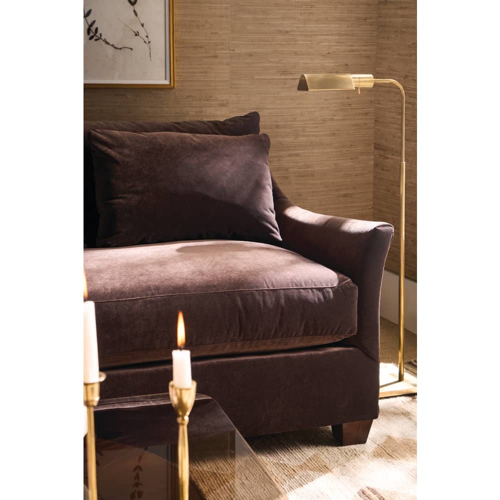 Brown velvet sofa with pillows next to a brass floor lamp in a warm, textured room.
