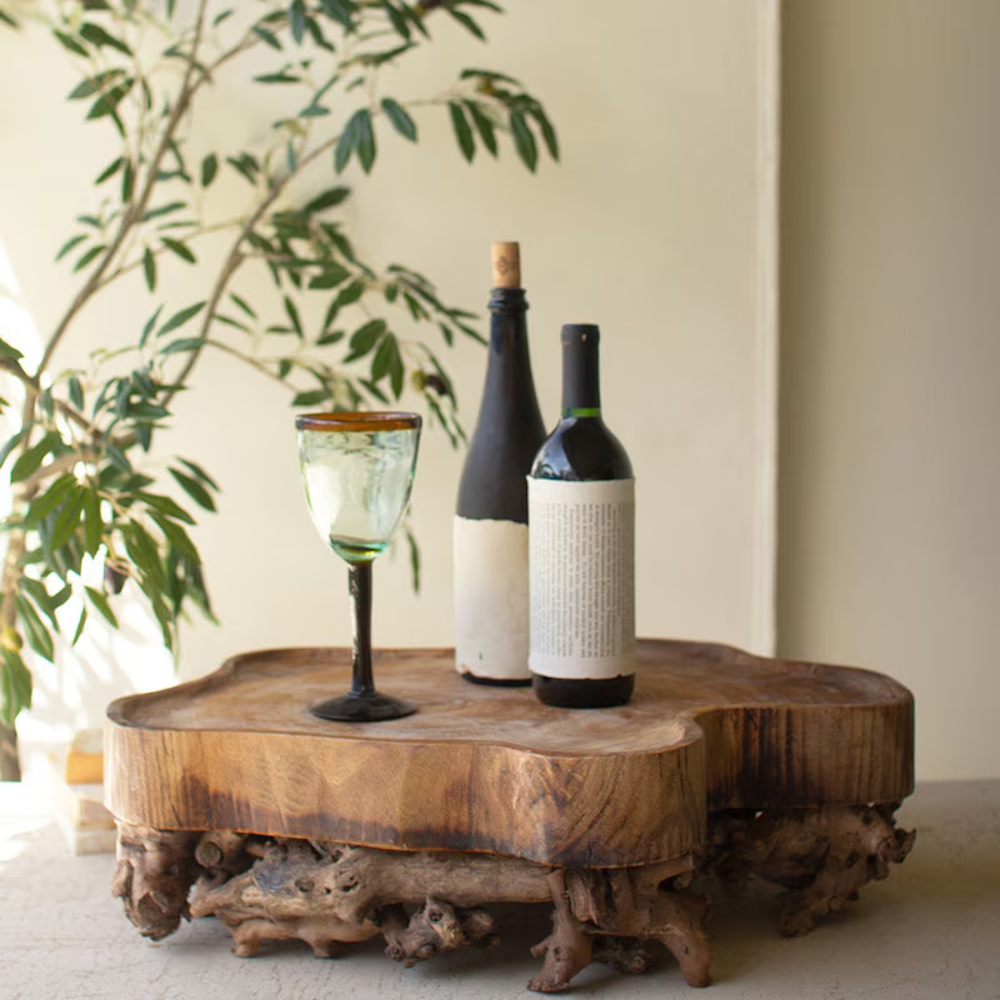 Rustic wooden slab tray with a gnarled root base, holding two wine bottles and a glass.