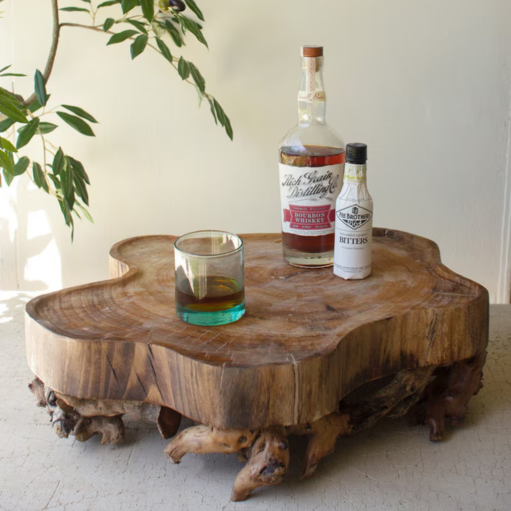 Organic-shaped wooden serving tray with a gnarled root base holding a glass, whiskey, and bitters.