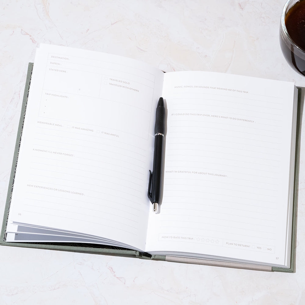 An open guided travel journal with lined prompts and a black pen resting on the pages next to a cup of coffee.