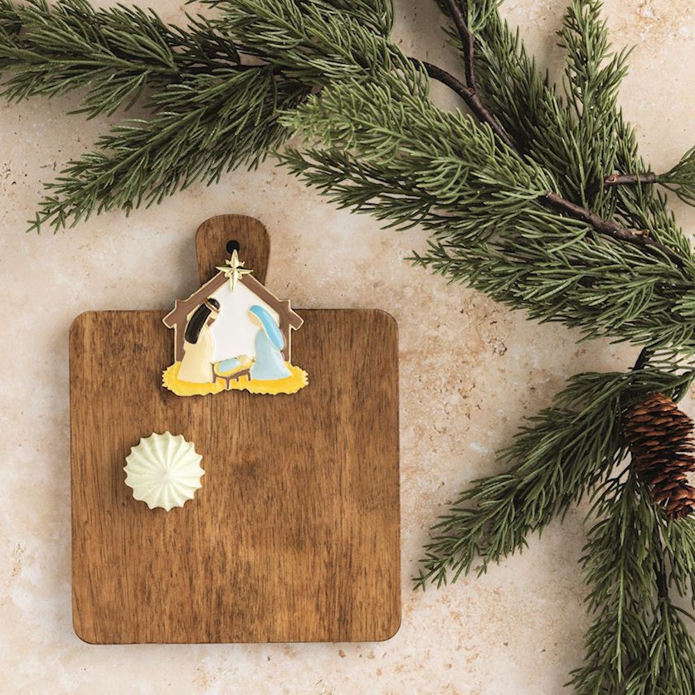 Nativity scene charm on a wooden cutting board next to a pine branch with a pinecone.