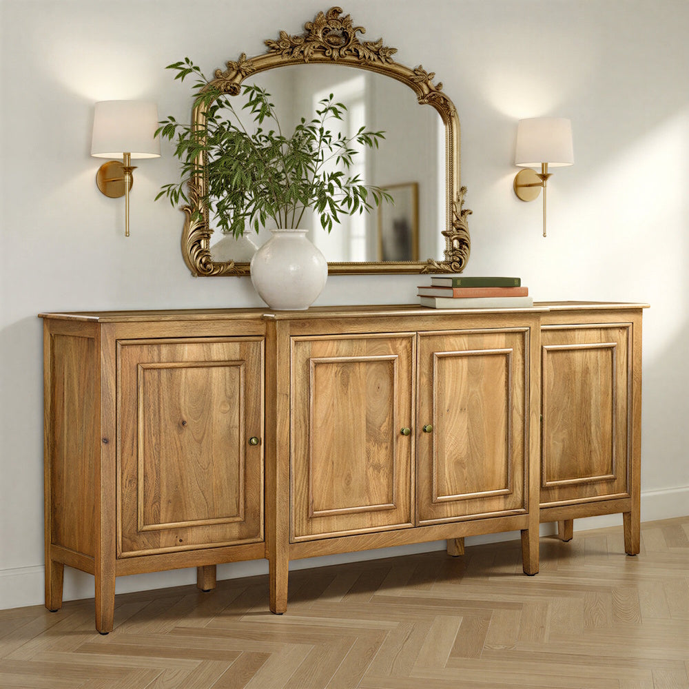 Natural hardwood four-door sideboard styled with a vase, books, ornate mirror, and wall sconces.