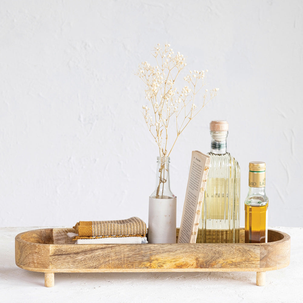 Long oval mango wood footed tray holding kitchen bottles, a vase with flowers, and linens.