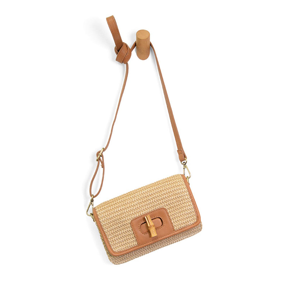 Natural straw crossbody bag with tan leather trim, bamboo turnlock closure, and adjustable strap hanging on a wooden peg.