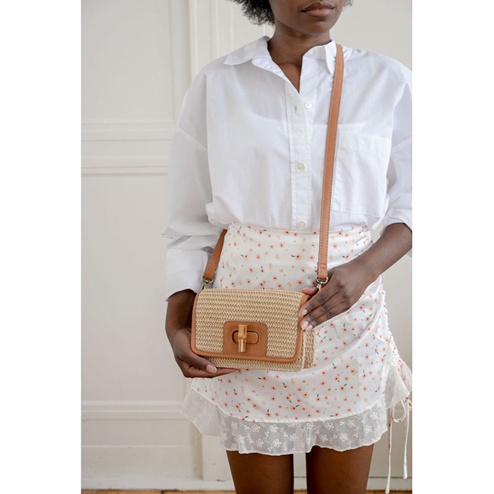 Model wearing a straw crossbody bag with bamboo turnlock over a white shirt and floral skirt.