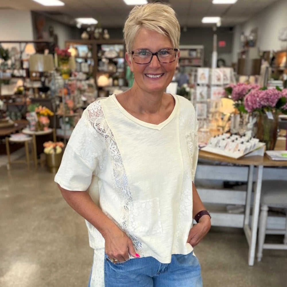 Blonde woman with glasses smiling in a boutique, wearing a cream patchwork lace v-neck top and blue jeans.