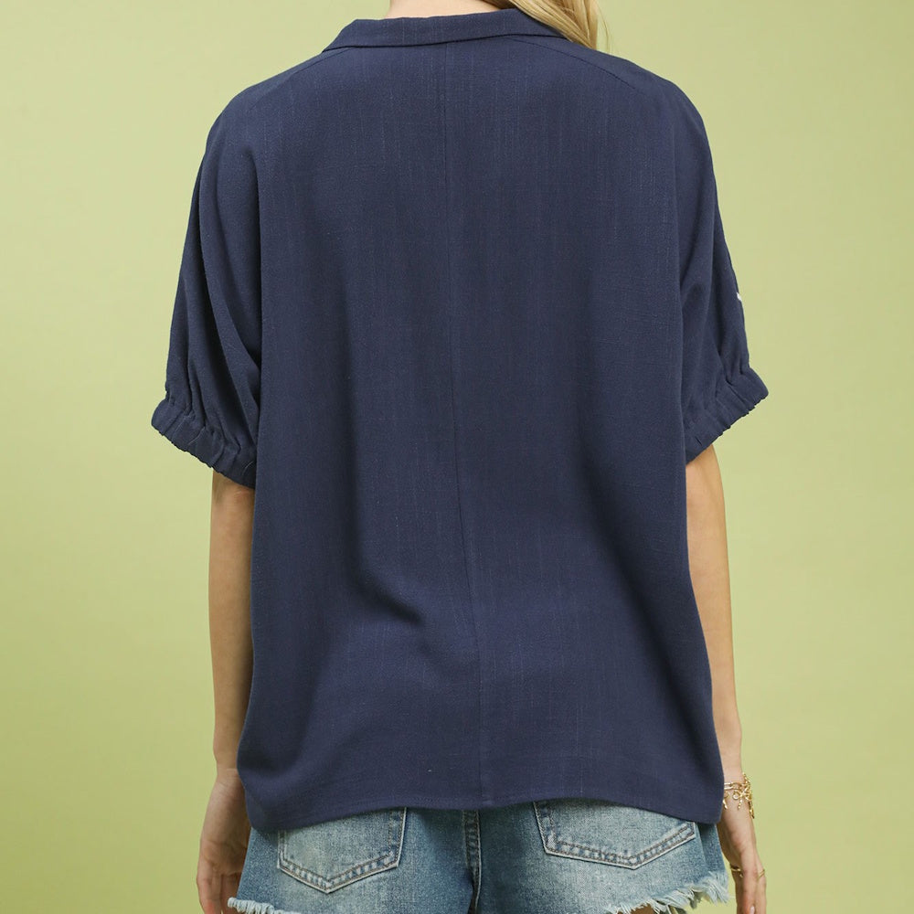 Back view of a woman wearing a loose navy blue short sleeve button down shirt with denim shorts.