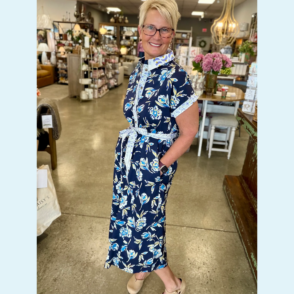 Woman in a navy floral midi shirt dress with a tie belt and button front, standing in a boutique.