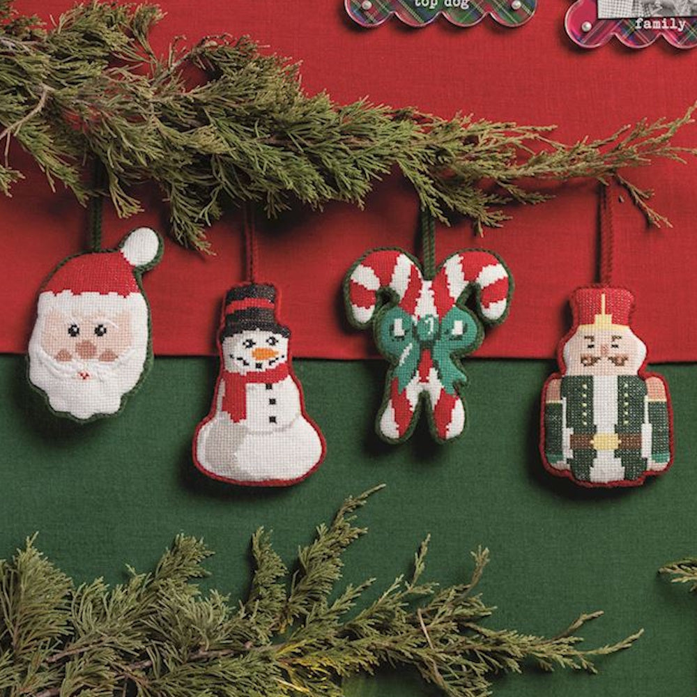 Four needlepoint Christmas ornaments: Santa, snowman, candy canes, and a nutcracker hanging on pine.