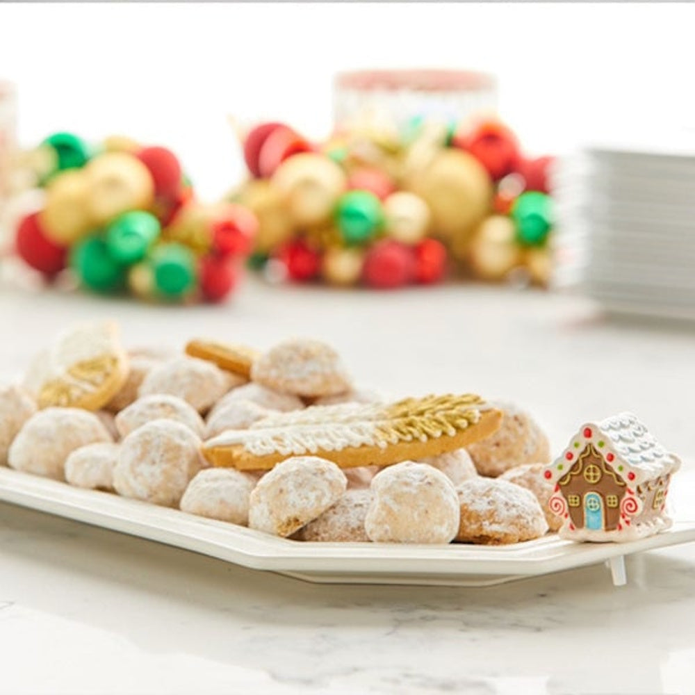 A ceramic gingerbread house on the edge of a white platter filled with powdered sugar cookies.