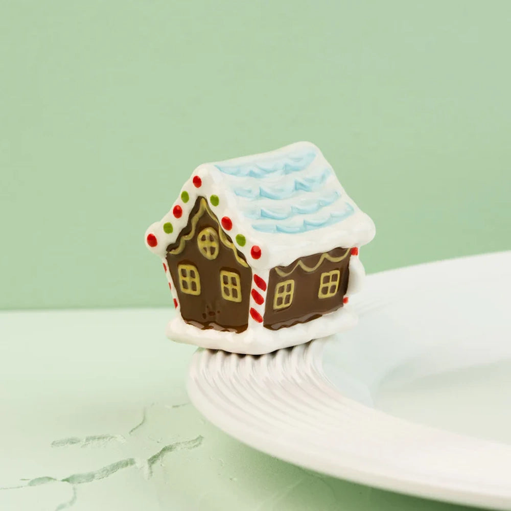 Mini ceramic gingerbread house figurine with candy decorations, sitting on the rim of a white plate.