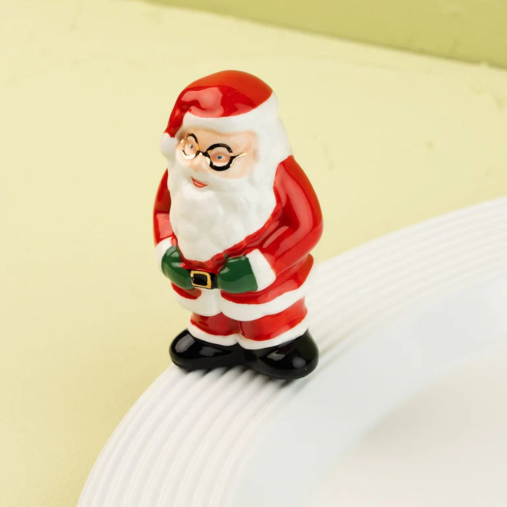 A miniature ceramic Santa Claus figurine wearing glasses, perched on the rim of a white serving platter.