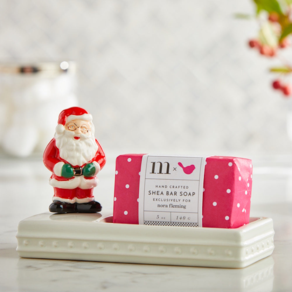 A ceramic Santa Claus figurine next to a pink wrapped bar of soap on a white ceramic soap dish.