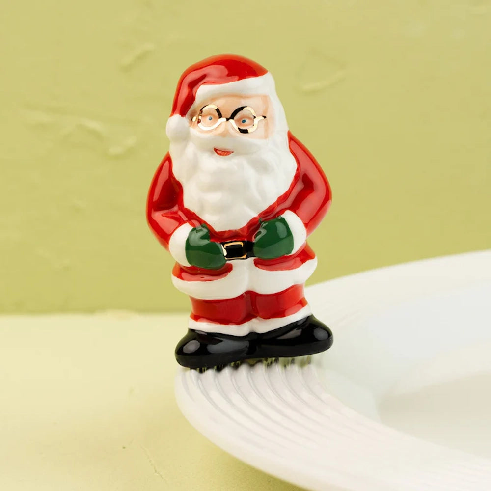 Ceramic Santa Claus figurine wearing glasses and green mittens, standing on the rim of a white plate.
