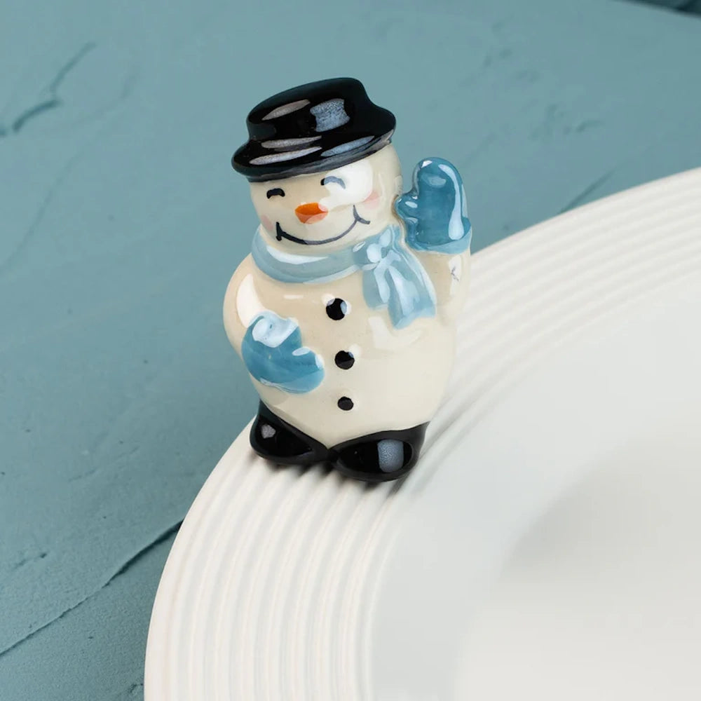 Small ceramic snowman figurine with a black top hat and blue scarf, waving from the edge of a white plate.