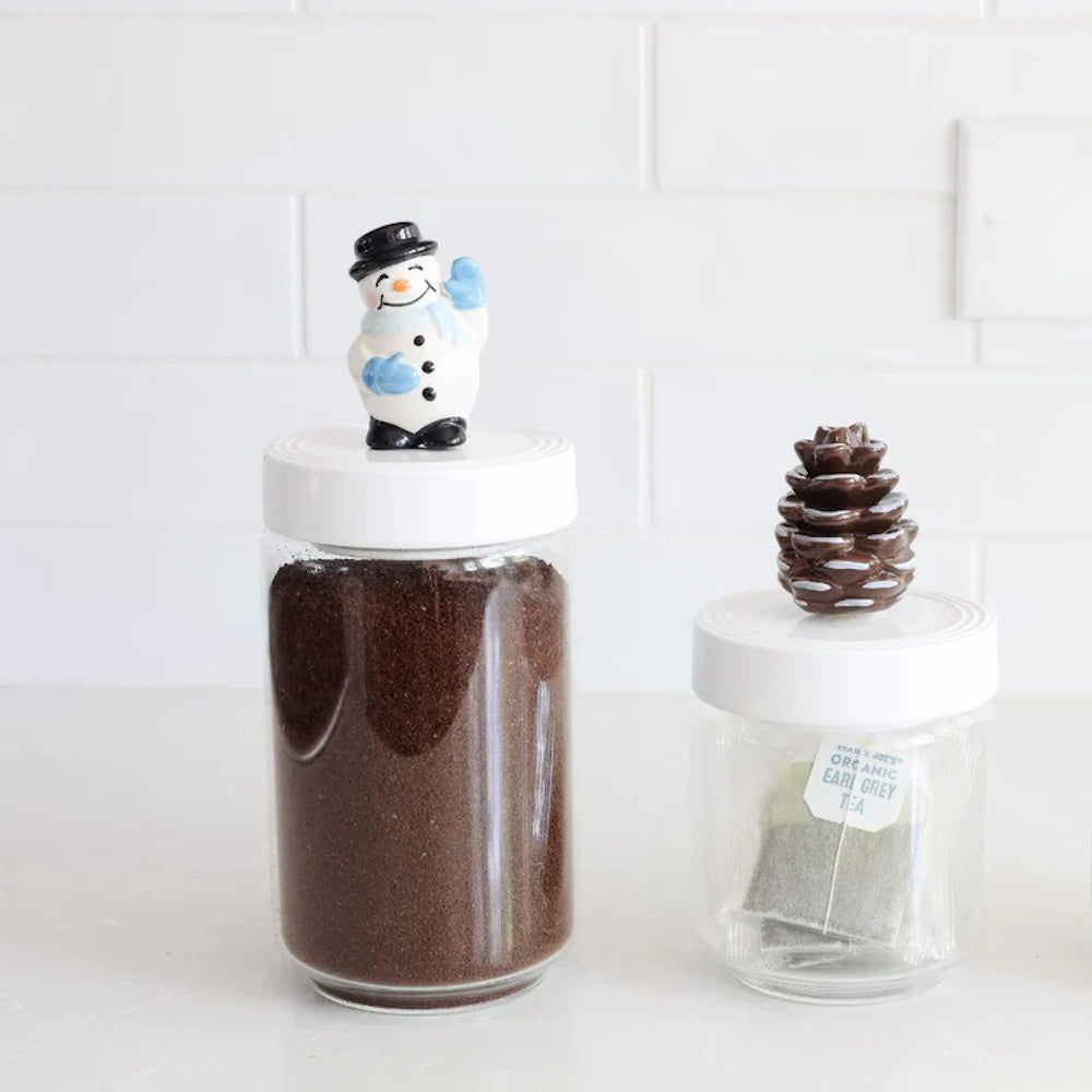 Two glass jars with white lids, one topped with a small snowman and the other with a brown pinecone figurine.