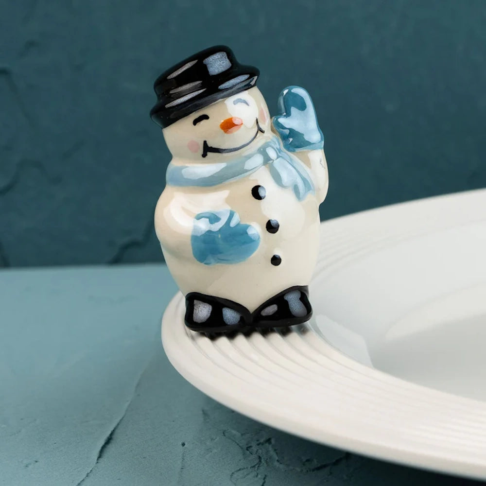 Ceramic snowman figurine wearing a black hat and blue scarf, waving while sitting on the edge of a white plate.