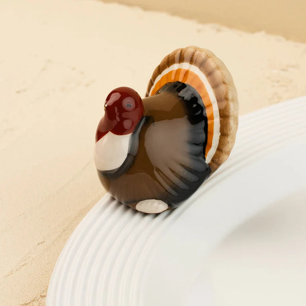 Small ceramic turkey figurine with an orange and brown fanned tail on a white plate rim.