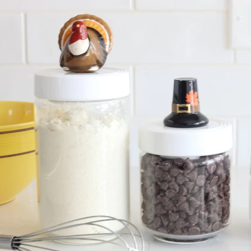 Two glass kitchen canisters with Thanksgiving-themed lid toppers, one holding flour and one chocolate chips.
