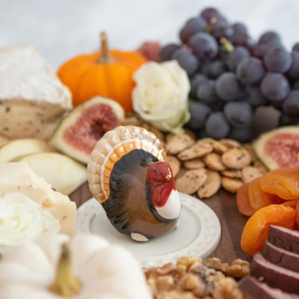 Ceramic turkey figurine on a charcuterie board with grapes, figs, pumpkins, cheese, and nuts.