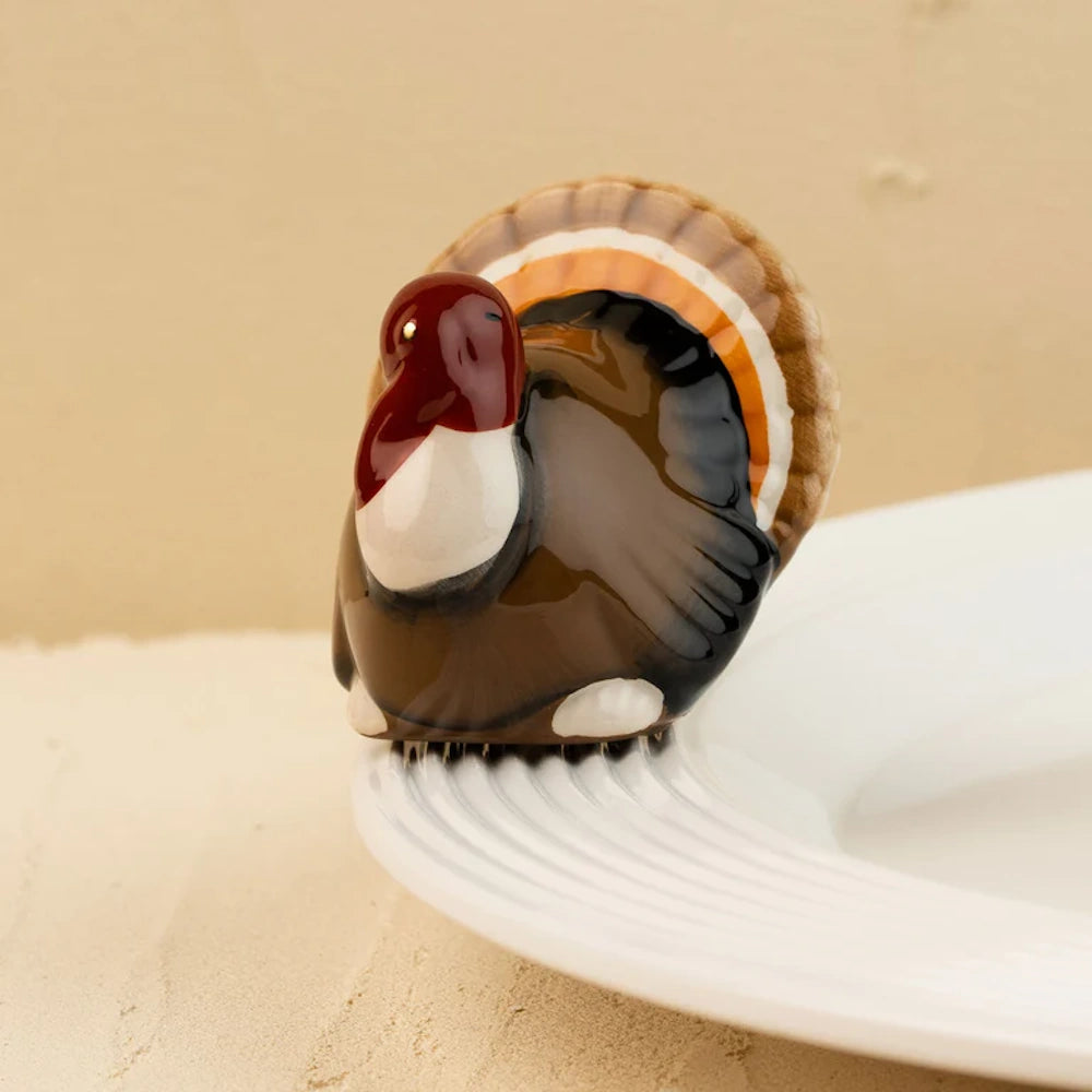 A small, glossy ceramic turkey figurine perched on the rim of a white plate.