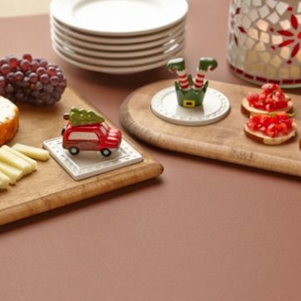 Holiday appetizers on wooden boards with a tiny red car and elf feet figurines next to a stack of plates.