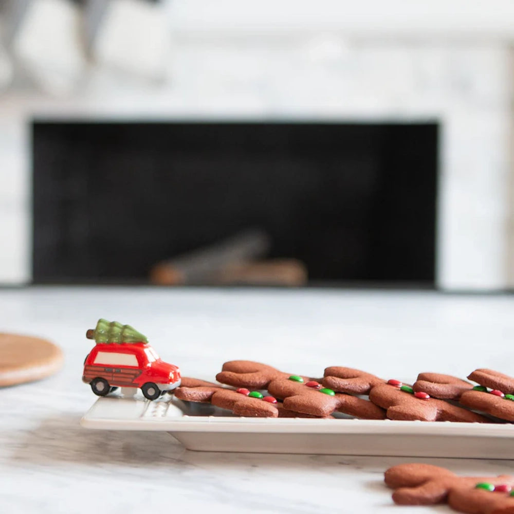 Gingerbread man cookies on a white platter decorated with a small red car carrying a Christmas tree.