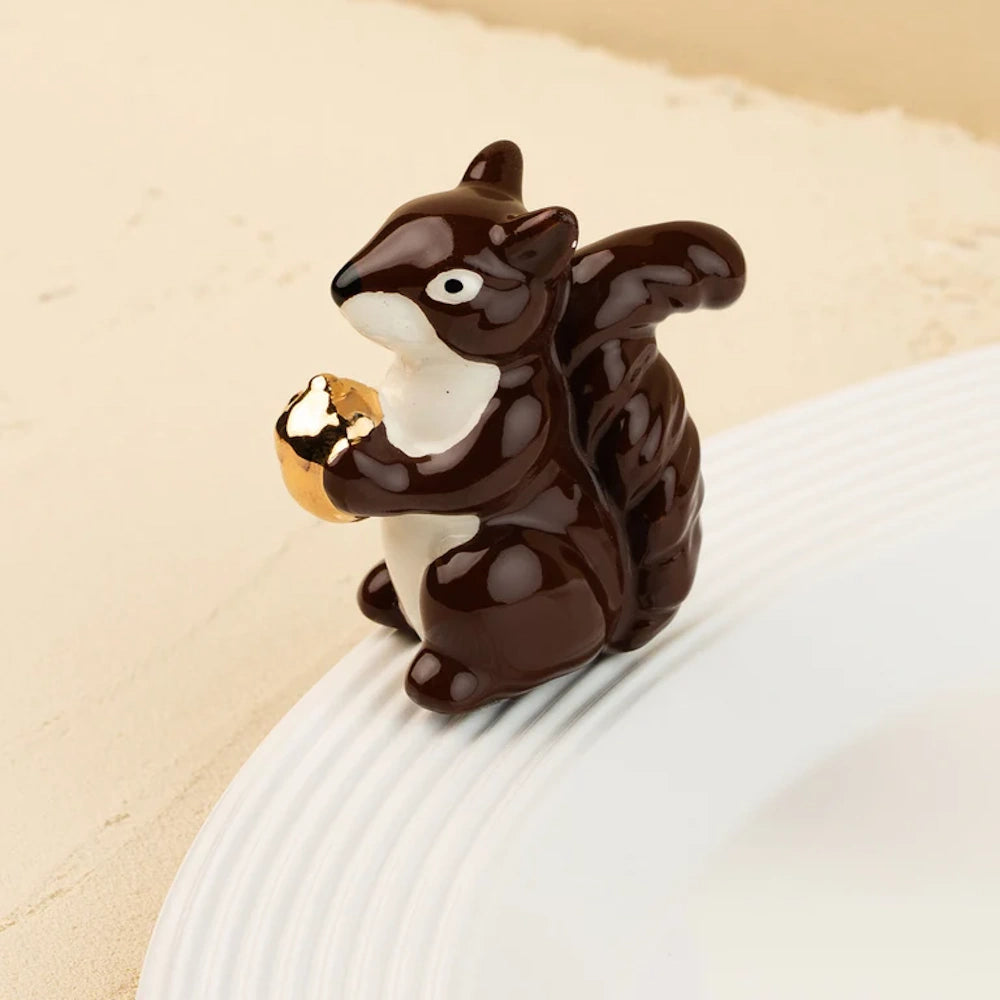 A small ceramic dark brown squirrel figurine with a white belly, holding a shiny gold acorn on a white plate.