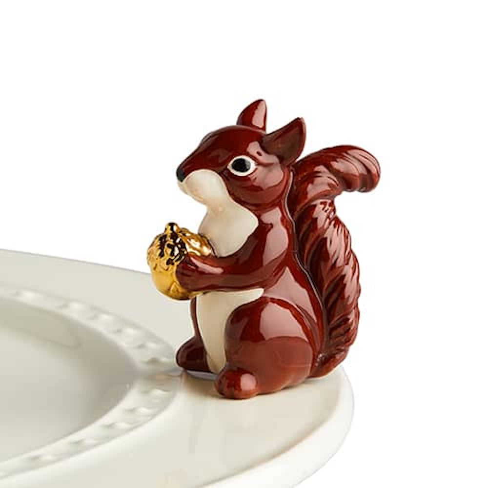 A glossy, brown ceramic squirrel figurine holding a golden acorn, attached to the rim of a white plate.