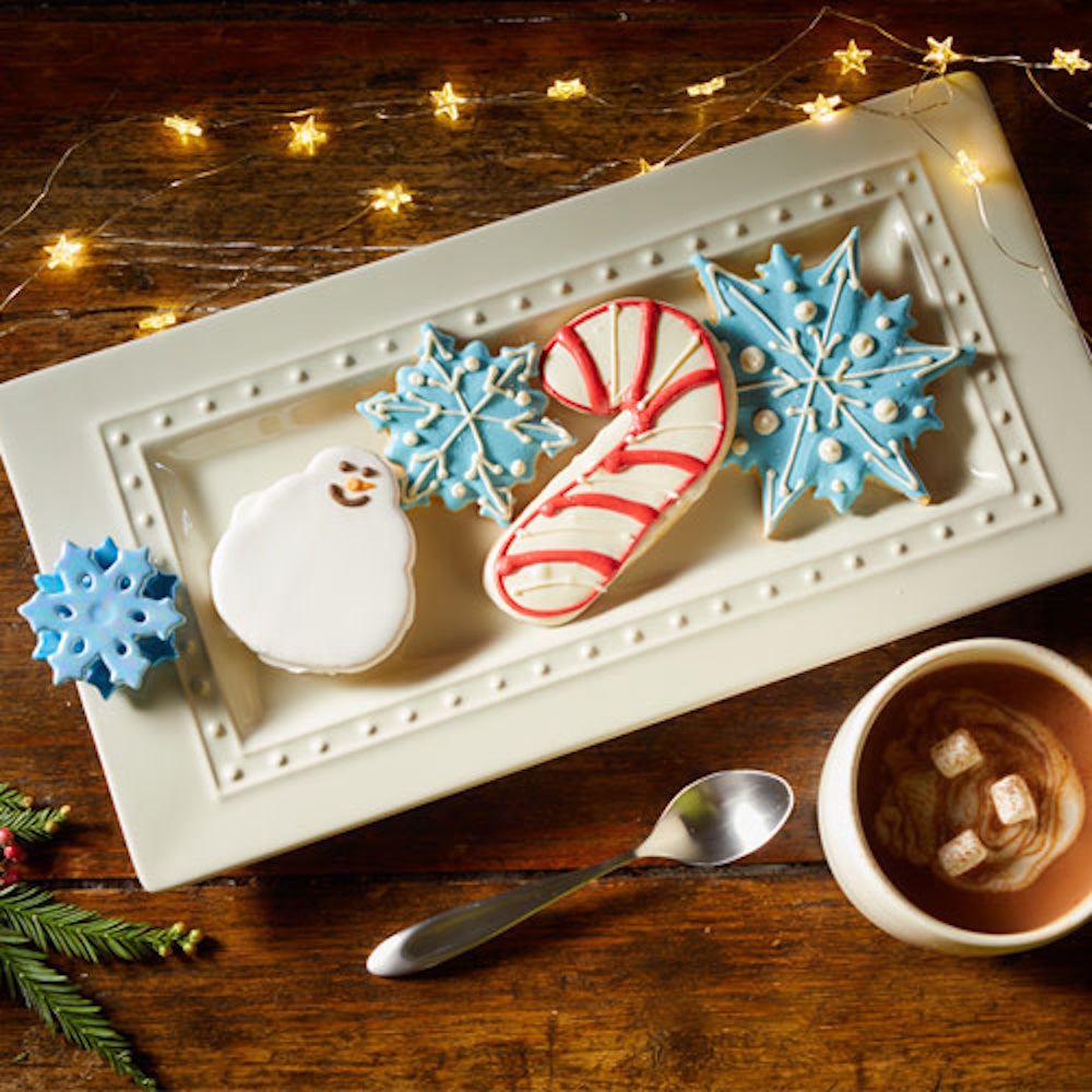 Christmas cookies on a platter next to a mug of hot cocoa, string lights, and a small blue snowflake charm.