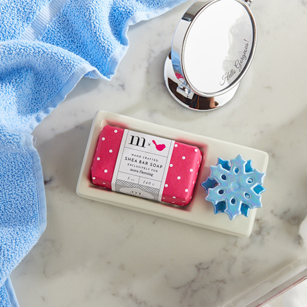 A wrapped pink bar soap and a blue snowflake charm in a white ceramic dish next to a blue towel and mirror.