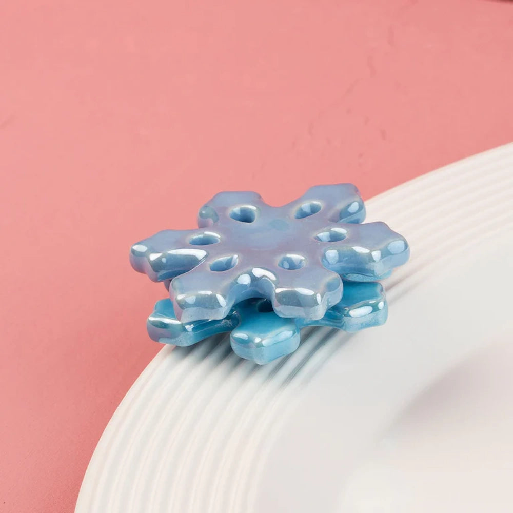 Iridescent ceramic blue snowflake charm resting on the rim of a white plate against a pink background.