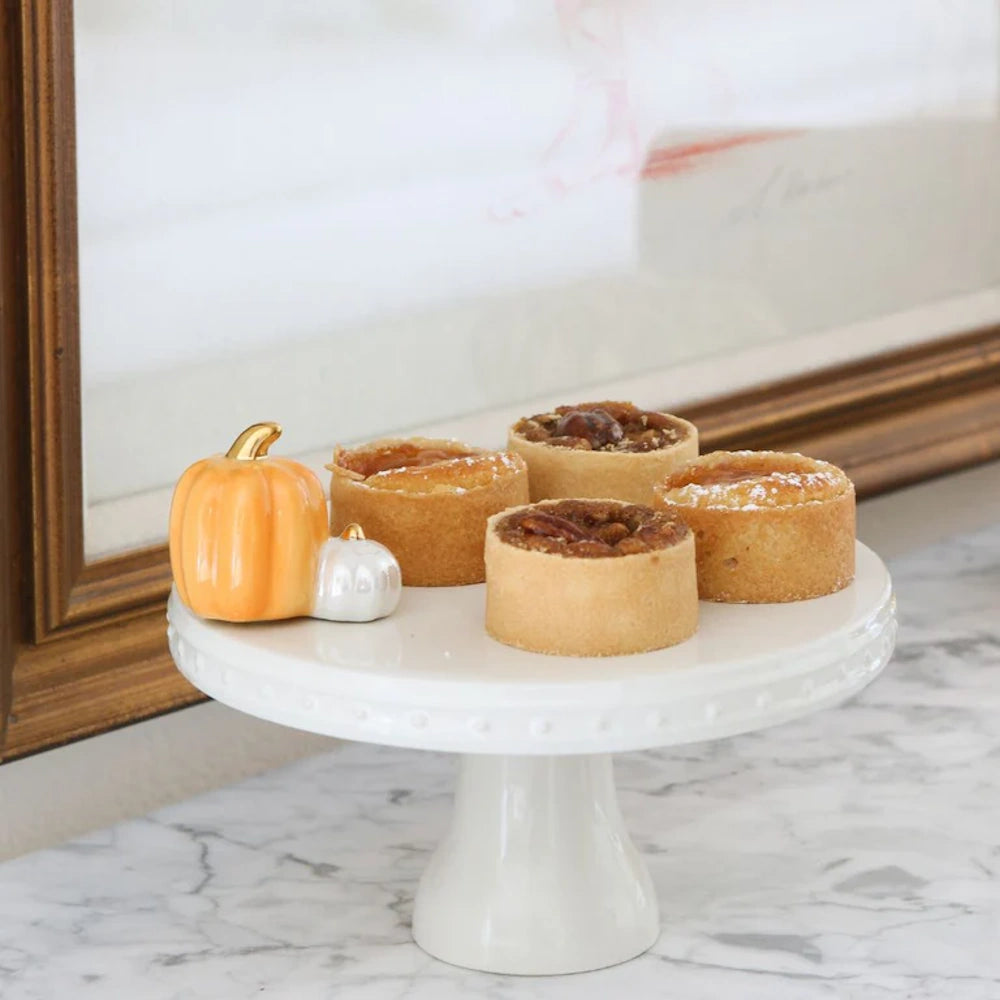 A miniature ceramic pumpkin figurine sits next to four small dessert tarts on a white cake stand.