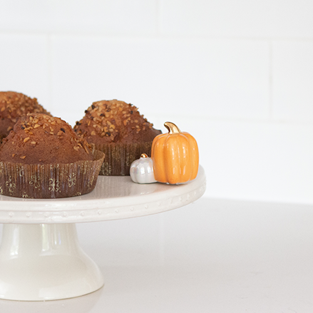 An orange and a white ceramic pumpkin figurine sitting on a cake stand next to three baked muffins.