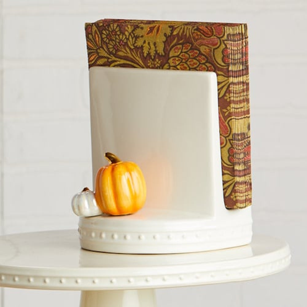 Ceramic pumpkin figurines attached to a white ceramic napkin holder holding patterned fall napkins.