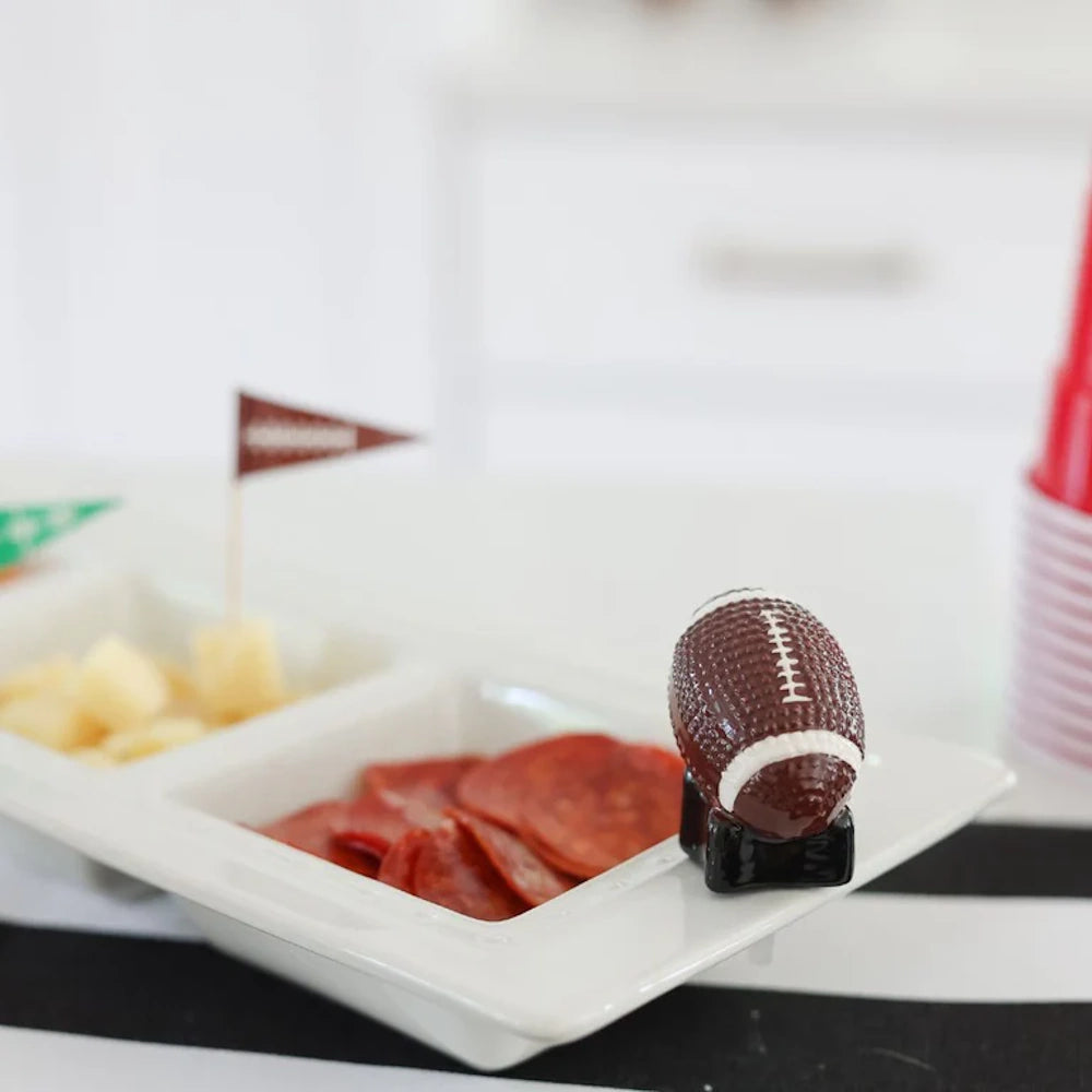 A miniature ceramic football figurine stands on the corner of a white divided tray holding meat and cheese.