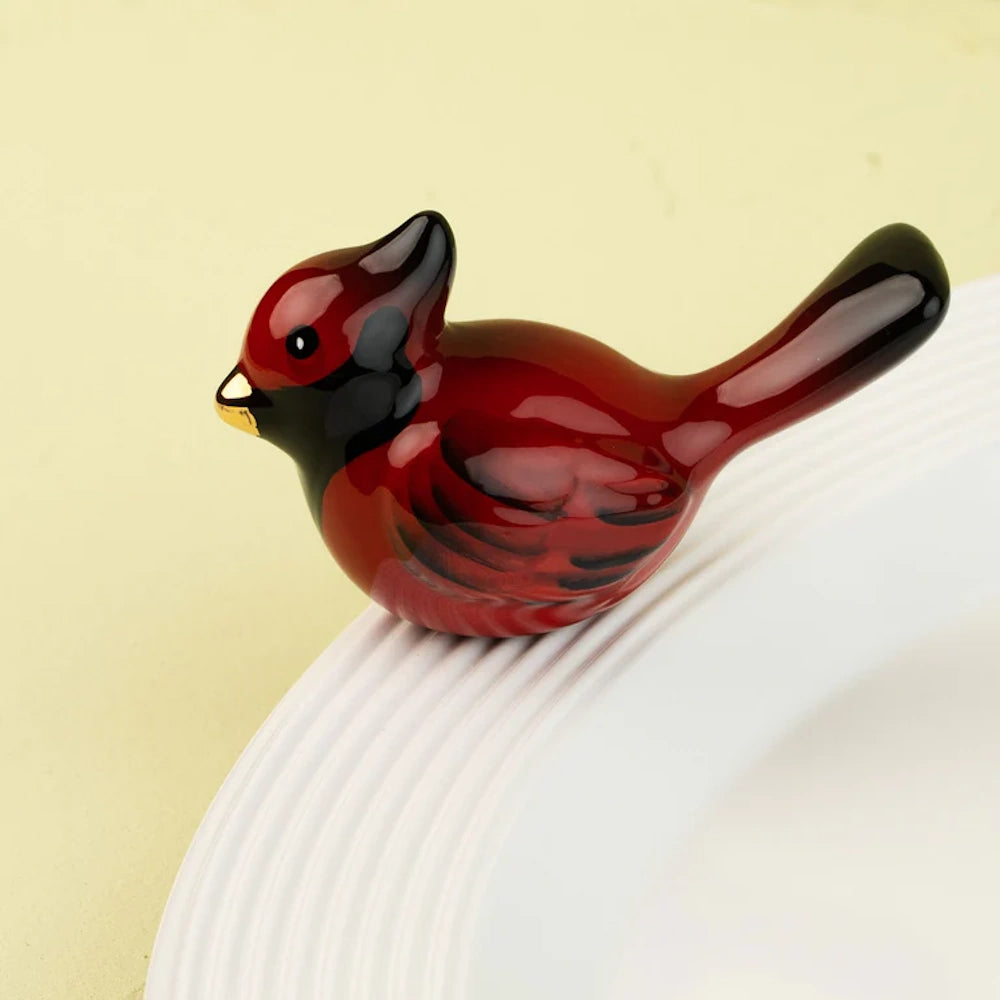 A glossy red ceramic cardinal figurine with black details and a gold beak, perched on a white plate rim.