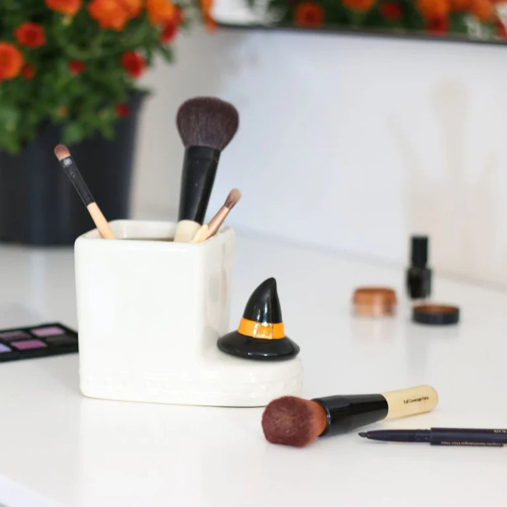 White ceramic makeup brush holder decorated with a small black witch hat, surrounded by cosmetics.