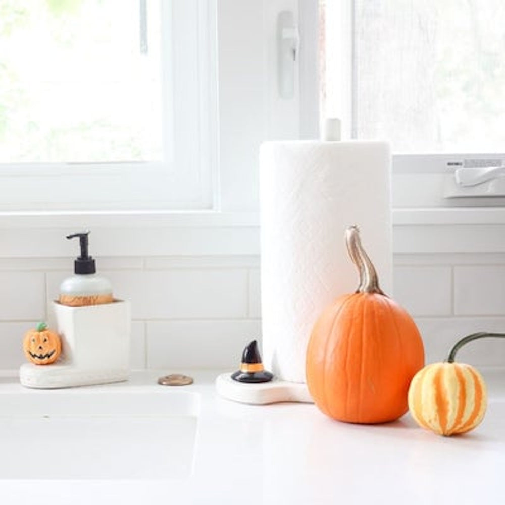 Halloween kitchen decor with pumpkins, a tiny witch hat, and a soap dispenser by a bright window.