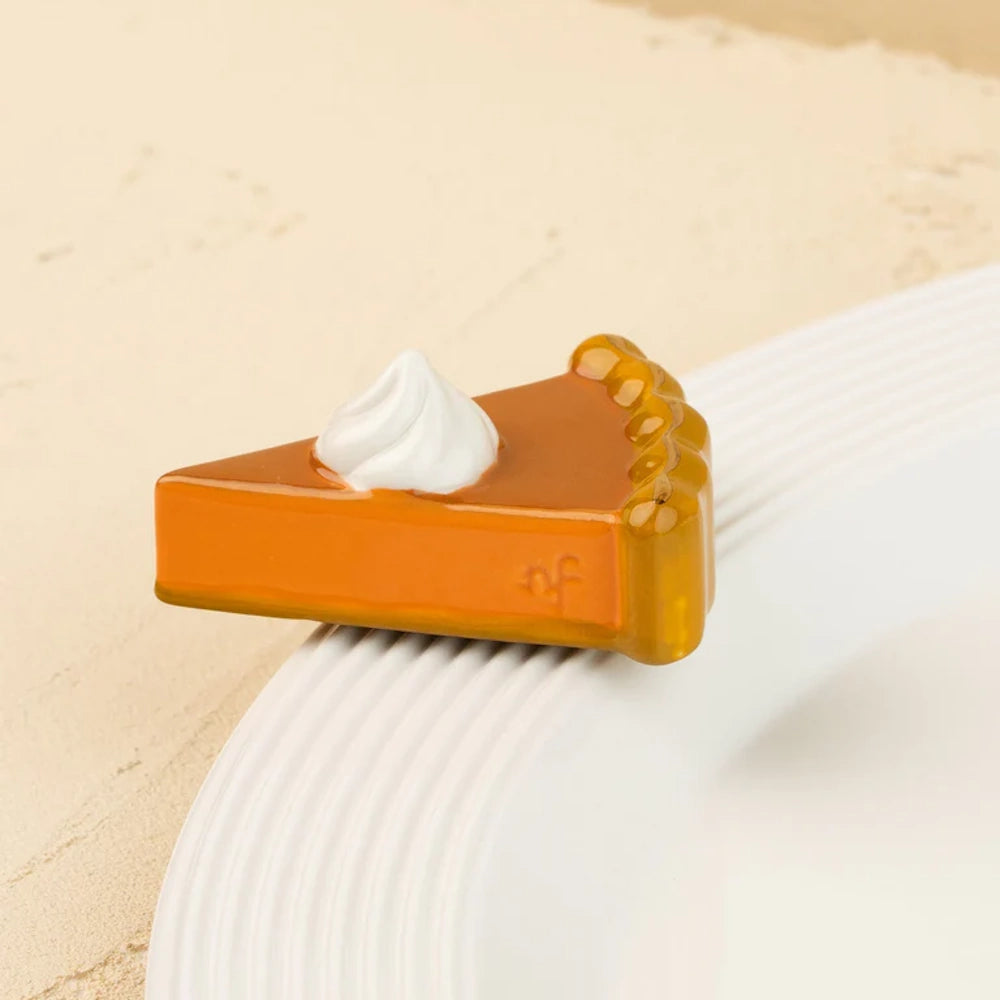A small ceramic slice of pumpkin pie with whipped cream, attached to the rim of a white plate.