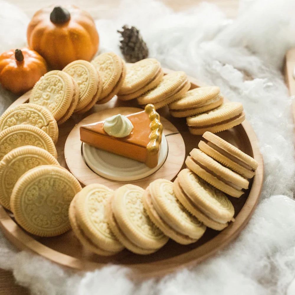 Ceramic pumpkin pie slice figurine on a wooden tray surrounded by sandwich cookies and mini pumpkins.