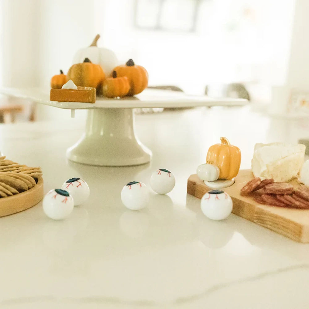 A bright autumn table spread with a mini pie slice, pumpkins on a pedestal, cheese, crackers, and toy eyeballs.