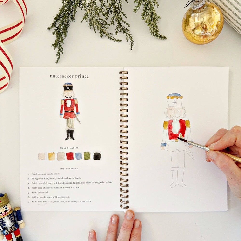 Hand using a paintbrush to color a Nutcracker prince in a spiral-bound watercolor workbook.