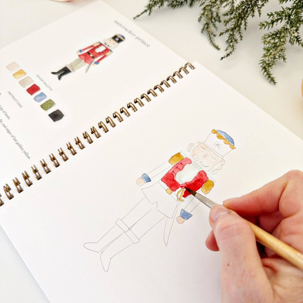 Close-up of a hand using a fine paintbrush to detail a red Nutcracker prince sketch in a workbook.