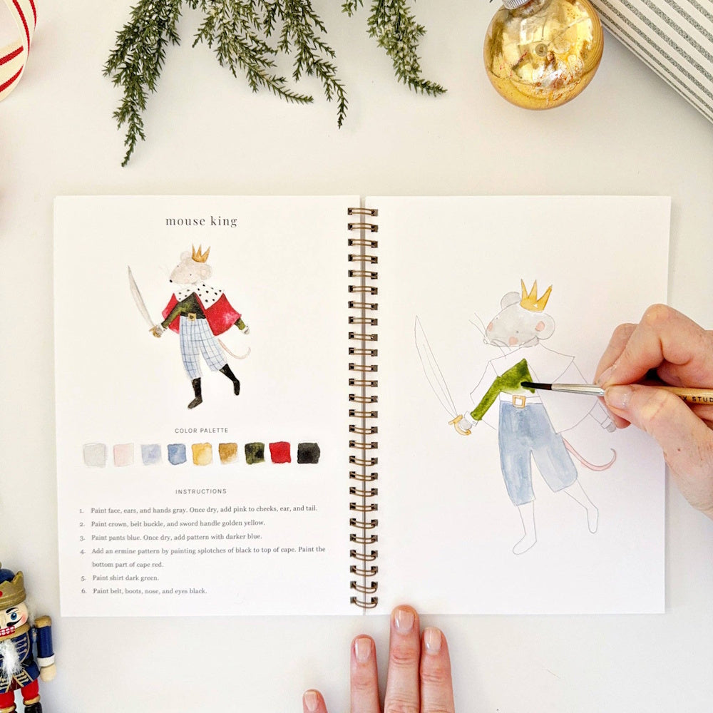 Hand painting a green sleeve on a Mouse King illustration in an open watercolor workbook.