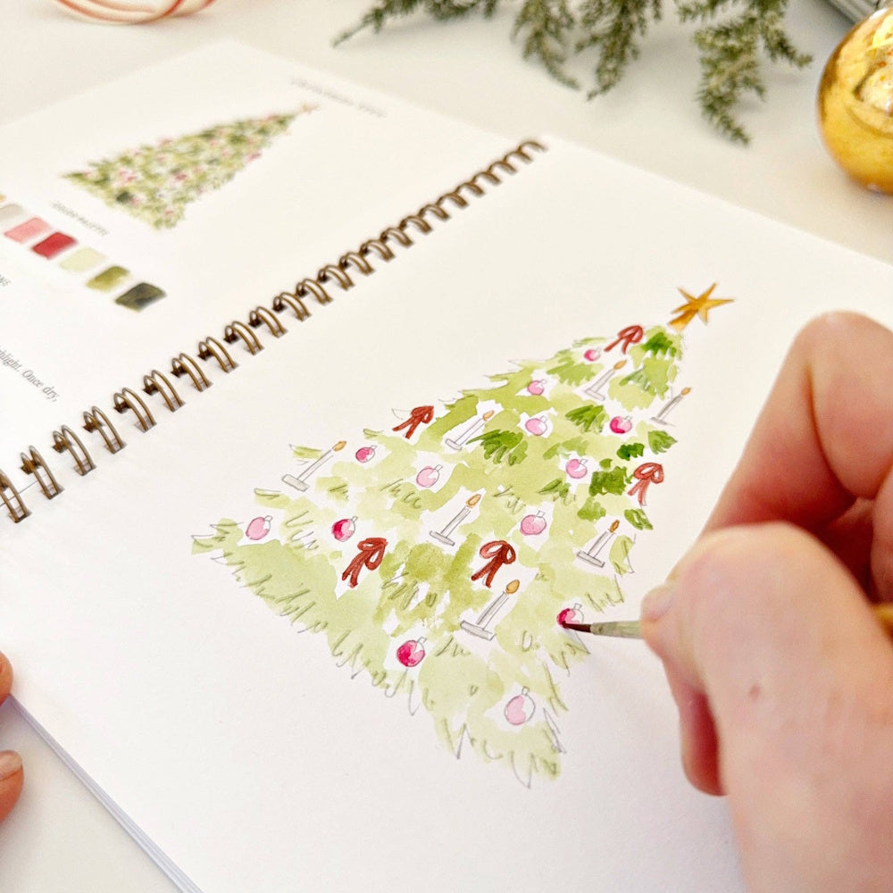Close-up of a hand adding red details to a watercolor Christmas tree illustration in a workbook.
