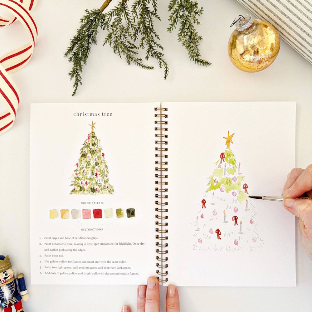 Hand using a paintbrush to color a Christmas tree sketch in an open watercolor workbook.