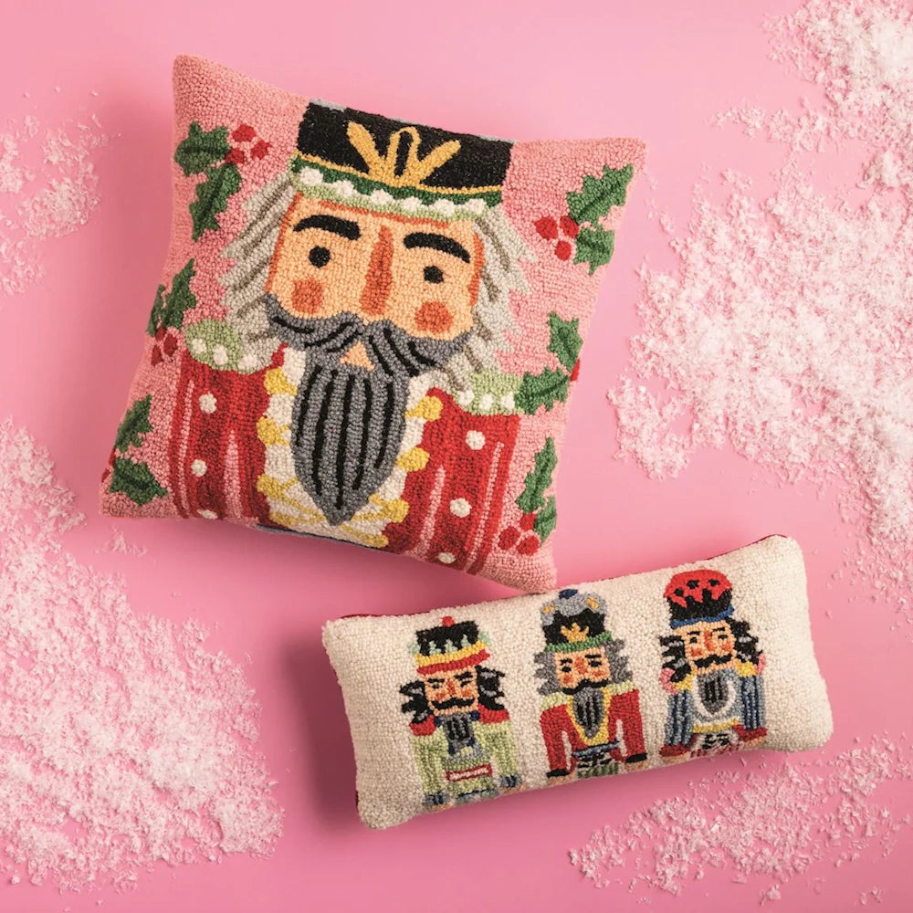 Two wool hook pillows featuring nutcracker designs on a pink background with artificial snow.