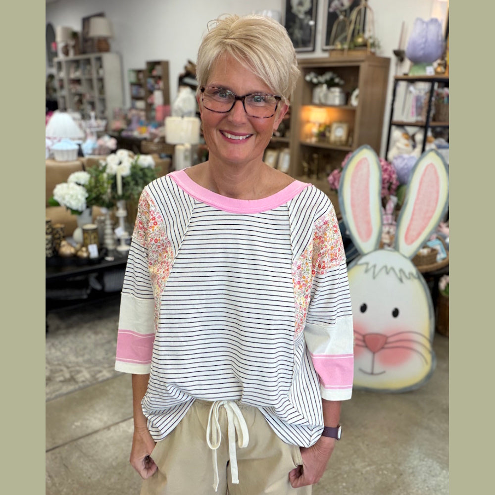 A woman with short blonde hair and glasses smiles in a boutique, wearing a striped top with floral accents.