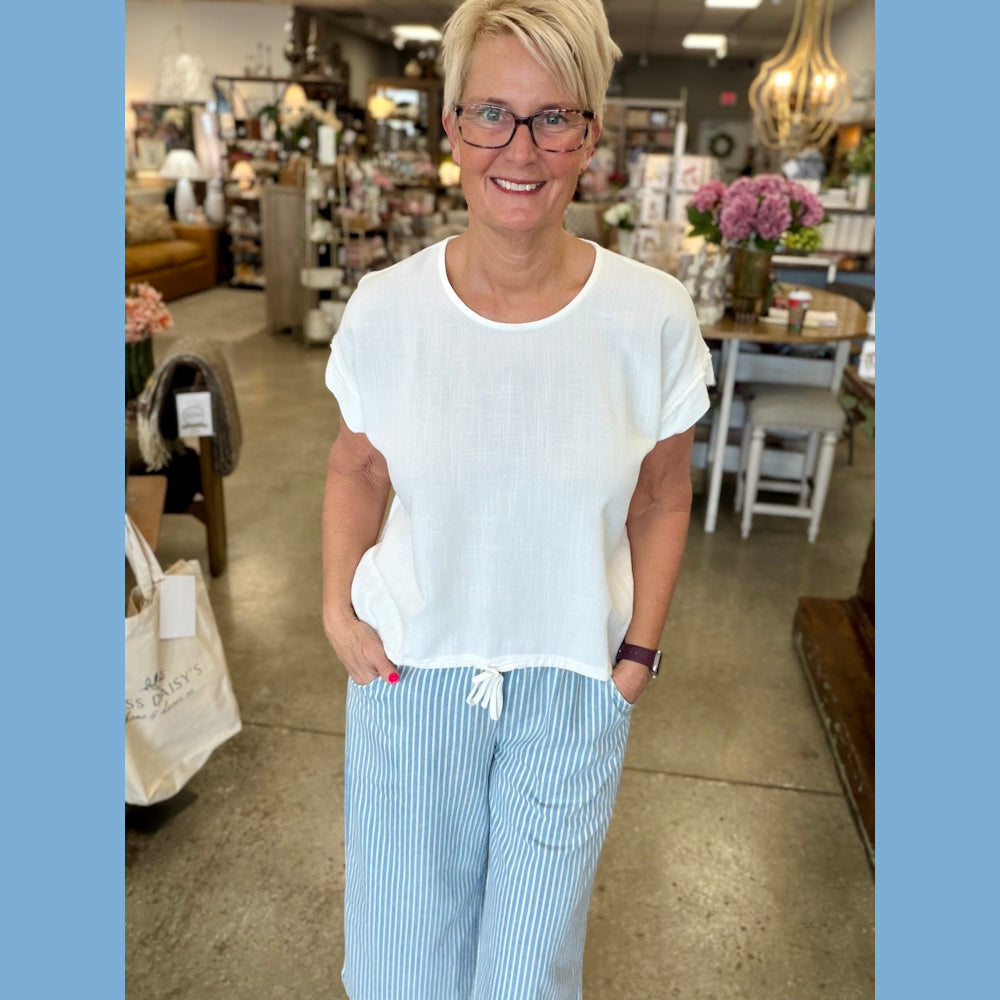 Woman wearing an off-white linen-blend drawstring top and blue striped pants in a boutique home decor store setting.